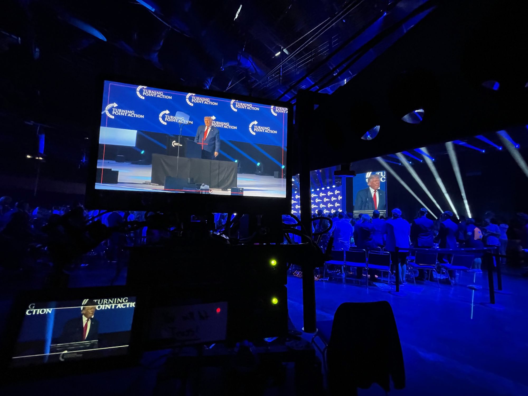 A speaker on a blue-lit stage shown large on the flown screen, with a confidence monitor and crew area in the dark foreground — Tampa, July 2022.