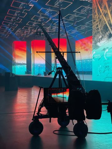 A camera rig on its wheeled base silhouetted against a wall of colour panels and an illuminated ceiling grid.
