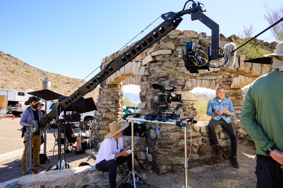 A jib-mounted camera reaching overhead on a desert location shoot, with a seated subject on a stone wall, a tripod camera nearby and crew standing around.