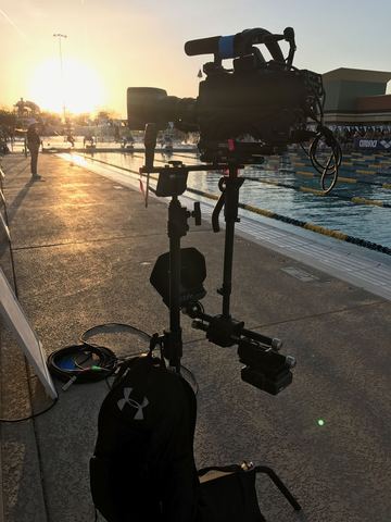 ESPN Swimming (2017) — a large video camera in silhouette against a low sun, with a blue pool, lane ropes and a concrete deck beyond.