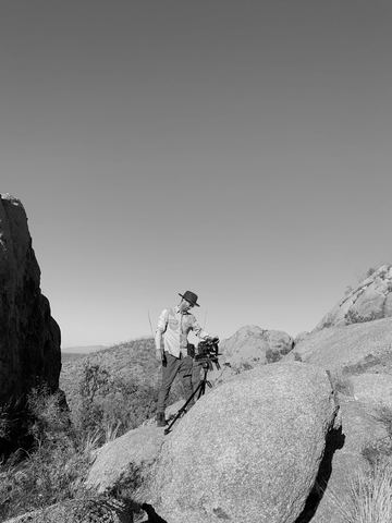 Camera Movement (2020) — a black-and-white frame of an operator in a wide-brimmed hat adjusting a tripod-mounted camera on a boulder, desert scrub and open sky behind.
